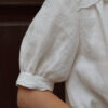 Lydia blouse sleeve slovakmeadows Woman wearing a light linen blouse with a V-neckline, collar, and lace details on the front, elbow-length sleeves.